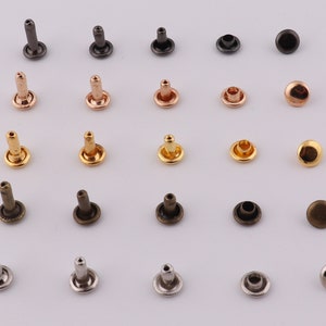 Op de afbeelding: Assortiment van metalen klinknagels in verschillende kleuren, waaronder zilver, goud, brons en zwart. De klinknagels zijn gerangschikt in rijen op een witte achtergrond.