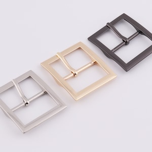 May include: Three metal belt buckles in different finishes: silver, gold, and black. The buckles are square shaped with a smooth, polished finish.
