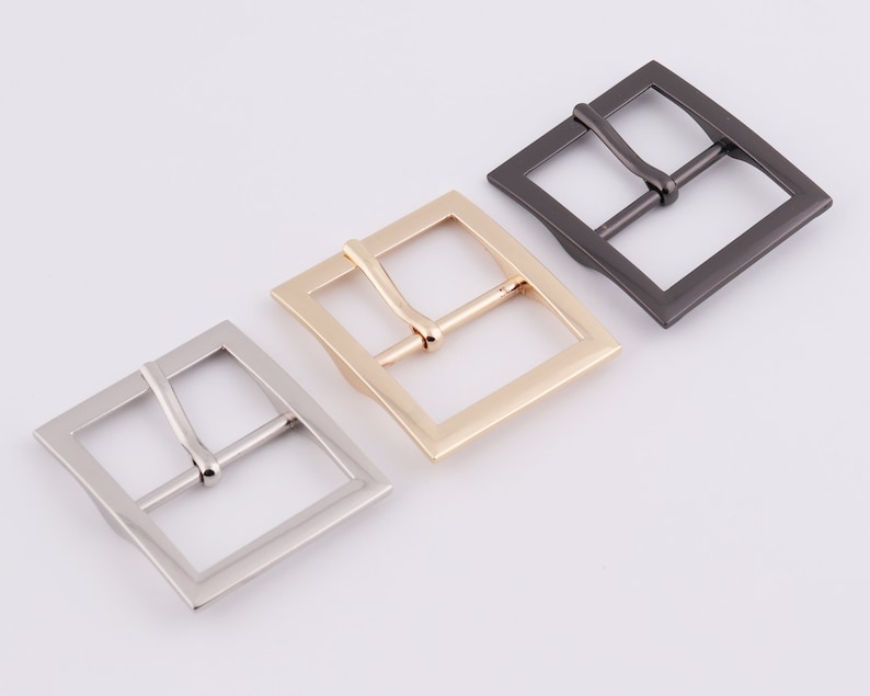May include: Three metal belt buckles in different finishes: silver, gold, and black. The buckles are square shaped with a smooth, polished finish.
