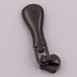 May include: A black metal zipper pull with a decorative design. The pull has a rounded top and a heart-shaped bottom.