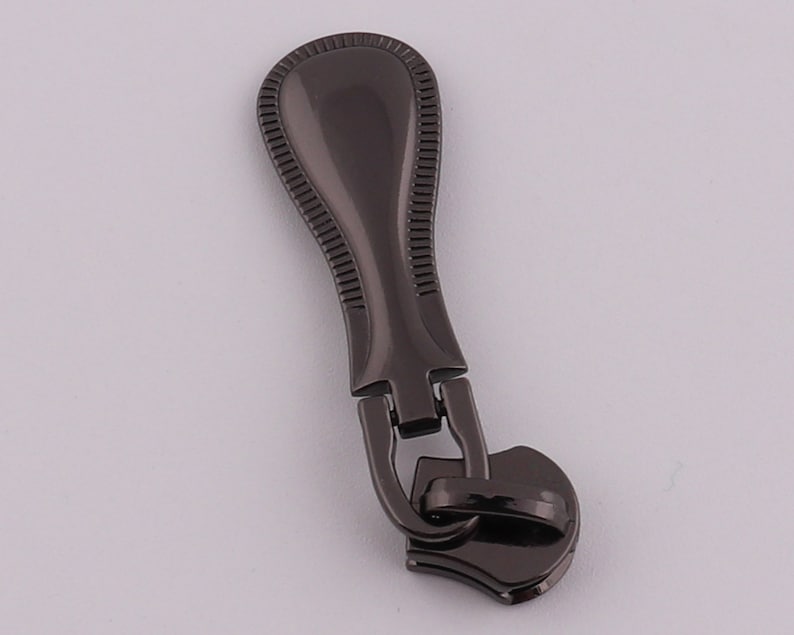 May include: A black metal zipper pull with a decorative design. The pull has a rounded top and a heart-shaped bottom.