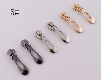 May include: Five zipper pulls in different colors: gold, silver, and black. The pulls are all the same shape, with a long, narrow body and a rounded end. They have a small hole at the top for attaching to a zipper.