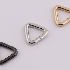 Op de afbeelding: Drie driehoekige metalen ringen in zwart, zilver en goud. De ringen zijn aan één kant open en hebben een gladde, gepolijste afwerking.