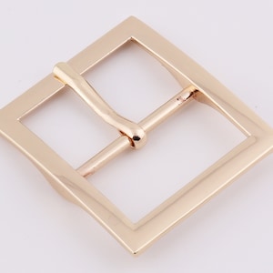 May include: A gold-toned metal belt buckle with a square frame and a smooth, polished finish.