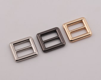 May include: Three metal square slides in silver, gunmetal, and gold colors. Each slide has two parallel bars across the center. The slides are likely used for adjusting straps on bags or other accessories.