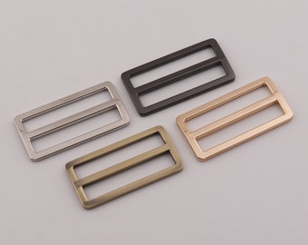 May include: Four metal slider buckles in different finishes: silver, black, gold, and antique brass. Each buckle has a rectangular shape with two parallel bars.