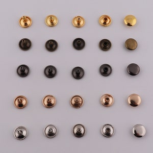 Peut inclure: Assortiment de 24 rivets métalliques de différentes couleurs, dont l'or, l'argent, le bronze et le noir. Les rivets sont disposés en rangées sur un fond blanc.