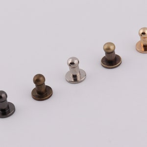 Op de afbeelding: Vijf metalen koepelvormige studs in verschillende afwerkingen: goud, messing, zilver, nikkel en zwart. De studs hebben een ronde basis en een koepelvormige bovenkant.