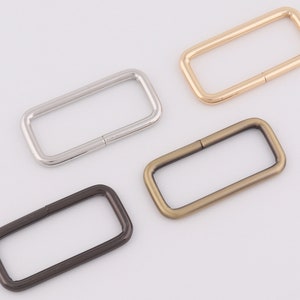 May include: Four rectangular metal rings in silver, gold, bronze, and black. Each ring has a split at one end, suggesting they are designed to be opened and closed. The rings are likely used for bag hardware or other crafting projects.