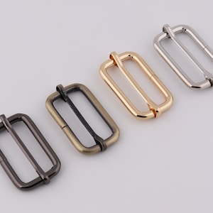 May include: Four metal slider buckles in different finishes: silver, gold, antique brass, and black. The buckles are rectangular with a center bar and are designed for attaching straps or belts.