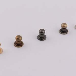 May include: Five metal dome-shaped push buttons with a flat base. The buttons are in different finishes: gold, silver, black, bronze, and brass.