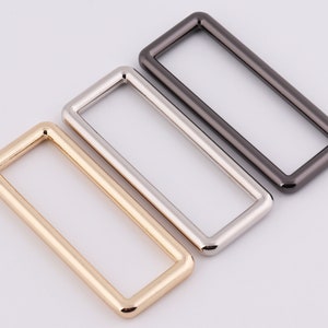 Könnte beinhalten: Drei rechteckige Metallringe in Gold, Silber und Schwarz.