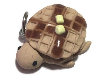 May include: A whimsical charm shaped like a turtle with a waffle shell, drizzled with syrup and topped with butter. The turtle has a light brown body and a silver-colored loop for attaching to a keychain or necklace.