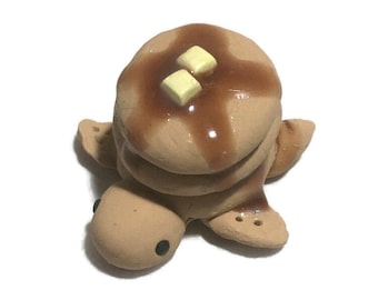 May include: A small figurine shaped like a turtle with a stack of three tan pancakes on its back. The pancakes are topped with a glossy brown syrup and two small yellow butter squares. The turtle has a tan body with two black dot eyes.