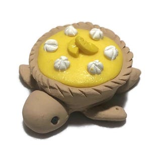 May include: A brown turtle figurine with a yellow lemon meringue pie on its back. The pie is decorated with white meringue swirls and lemon slices.