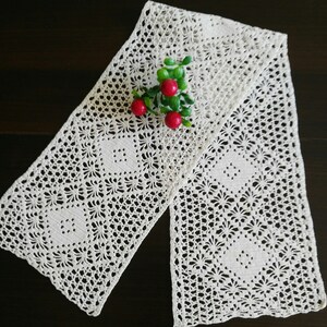 May include: A white crocheted table runner with a delicate floral pattern. The runner is folded in half and has a sprig of red berries on top.