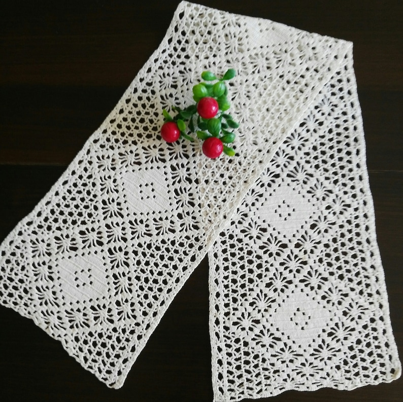 May include: A white crocheted table runner with a delicate floral pattern. The runner is folded in half and has a sprig of red berries on top.