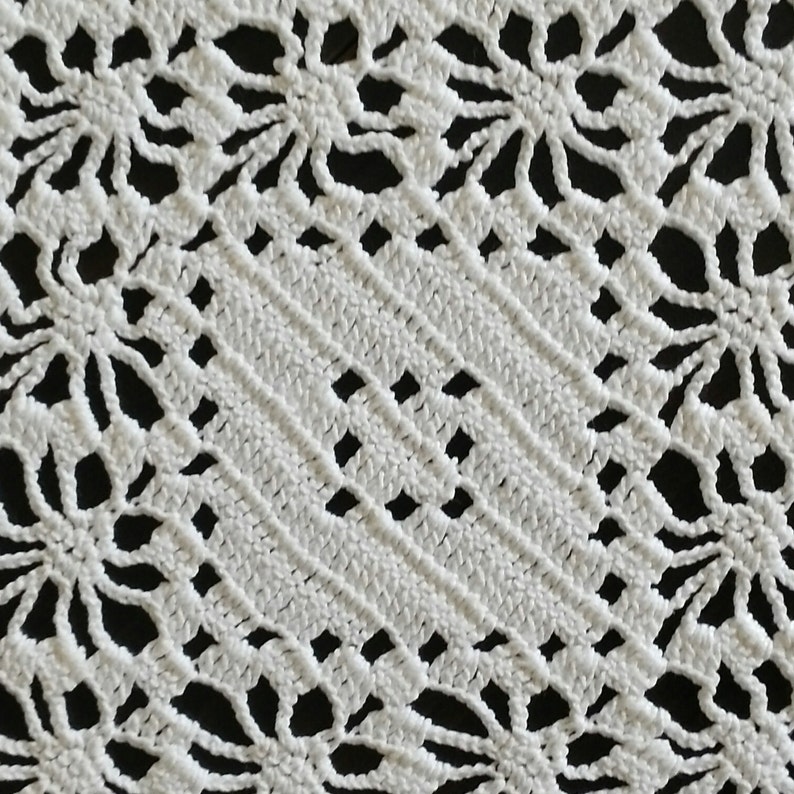 May include: White crocheted doily with a delicate floral pattern. The doily is square shaped with a central diamond design.