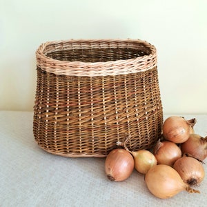 Puede incluir: Una cesta de mimbre tejida con un borde marrón claro, llena de cebollas marrones. La cesta está sobre una superficie de tela blanca.