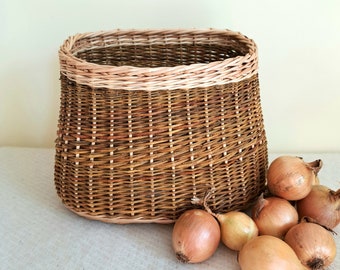 Puede incluir: Una cesta de mimbre tejida con un cuerpo marrón oscuro y un borde beige más claro, con forma rectangular redondeada. Varias cebollas amarillas enteras con pieles papiráceas están agrupadas cerca de la base, mostrando texturas y colores naturales.