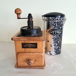 May include: A vintage wooden coffee grinder with a black metal mechanism and a drawer. The grinder has a label that reads "Braunag" and is next to a silver coffee canister with a black lid and a label that reads "Coffee".