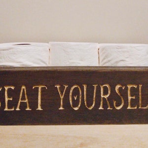 Peut inclure: Une bo&icirc;te en bois brun fonc&eacute; avec les mots "SEAT YOURSELF" grav&eacute;s sur le devant. La bo&icirc;te contient trois rouleaux de papier toilette blanc.
