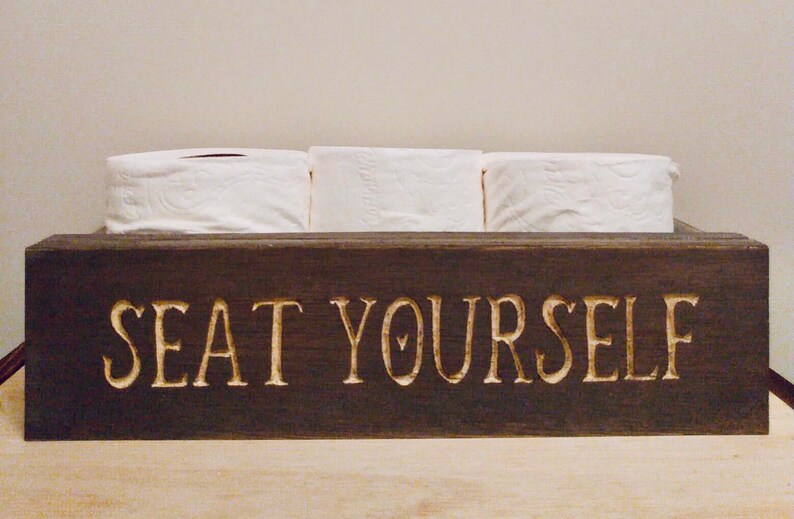 Peut inclure: Une bo&icirc;te en bois brun fonc&eacute; avec les mots "SEAT YOURSELF" grav&eacute;s sur le devant. La bo&icirc;te contient trois rouleaux de papier toilette blanc.