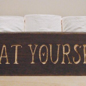 Peut inclure: Un porte-papier toilette en bois brun fonc&eacute; avec le texte "SEAT YOURSELF" grav&eacute; sur le devant. Trois rouleaux de papier toilette blanc sont pos&eacute;s sur le support.