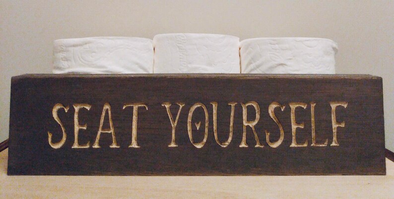 Peut inclure: Un porte-papier toilette en bois brun fonc&eacute; avec le texte "SEAT YOURSELF" grav&eacute; sur le devant. Trois rouleaux de papier toilette blanc sont pos&eacute;s sur le support.