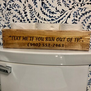 Peut inclure: Porte-papier toilette en bois avec le texte "Text me if you run out of TP" et le num&eacute;ro de t&eacute;l&eacute;phone (940) 552-7463.