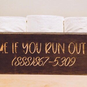 Peut inclure: Un porte-papier toilette en bois brun fonc&eacute; avec le texte "TEXT ME IF YOU RUN OUT OF TP (888)867-5309" grav&eacute; en lettres dor&eacute;es.