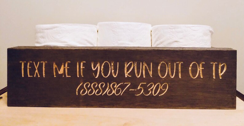 Peut inclure: Un porte-papier toilette en bois brun fonc&eacute; avec le texte "TEXT ME IF YOU RUN OUT OF TP (888)867-5309" grav&eacute; en lettres dor&eacute;es.