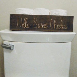 Peut inclure: Un porte-papier toilette en bois avec le texte "Hello Sweet Cheeks" en lettres dor&eacute;es. Le porte-papier est pos&eacute; sur un r&eacute;servoir de toilette blanc.