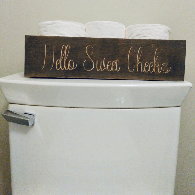 Peut inclure: Un porte-papier toilette en bois avec le texte "Hello Sweet Cheeks" en lettres dor&eacute;es. Le porte-papier est pos&eacute; sur un r&eacute;servoir de toilette blanc.