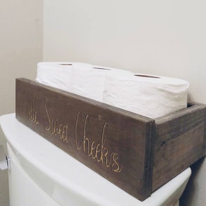Peut inclure: Un porte-papier toilette en bois rustique avec le texte "Hello Sweet Cheeks" grav&eacute; sur le devant. Le porte-papier toilette contient trois rouleaux de papier toilette.