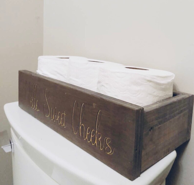 Peut inclure: Un porte-papier toilette en bois rustique avec le texte "Hello Sweet Cheeks" grav&eacute; sur le devant. Le porte-papier toilette contient trois rouleaux de papier toilette.