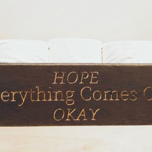 Peut inclure: Une bo&icirc;te en bois brun fonc&eacute; avec le texte "HOPE Everything Comes Out OKAY" grav&eacute; en lettres dor&eacute;es. La bo&icirc;te contient trois rouleaux de papier toilette blancs.