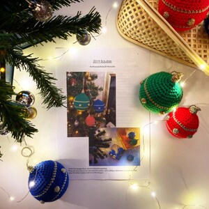 以下が含まれることがあります： 編み図のアップで、クリスマスオーナメントの作り方の説明が書かれています。赤、緑、青、白など、さまざまな色が使われています。編み図のタイトルは「2019 Baubles」です。