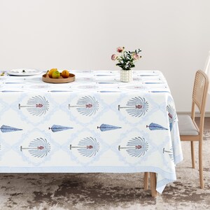 May include: A white tablecloth with a repeating blue and red floral pattern covers a table. The tablecloth has a blue striped border. A vase of pink flowers and a wooden tray with fruit sit on the table.