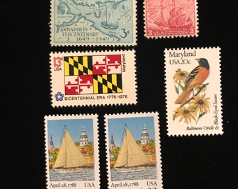 May include: A collection of six vintage postage stamps featuring the state of Maryland. The stamps depict a map of Annapolis, the Maryland state flag, a Baltimore Oriole bird, and two sailing ships. The stamps are from the 1970s and 1980s.