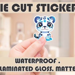 May include: Die cut sticker of a blue and white cartoon snow leopard. The sticker is being held in a hand. The text "Waterproof. Laminated Gloss. Matte" is at the bottom of the image.