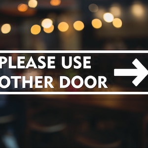 May include: White sign with black border and text that reads "PLEASE USE OTHER DOOR" with a white arrow pointing to the right.