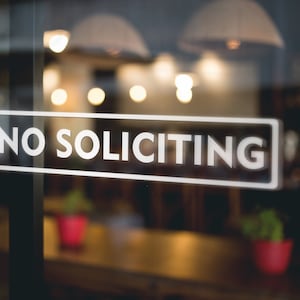 May include: A white sign with black lettering on a glass door reads "NO SOLICITING". The sign is in focus, while the background is blurred and shows a cafe interior with lights and plants.
