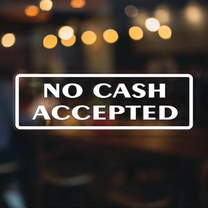 May include: A white sign with black text that reads "NO CASH ACCEPTED" on a blurred background of a cafe or restaurant.