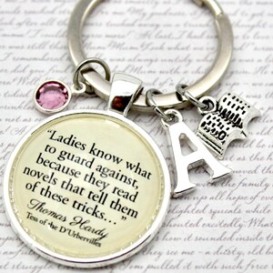 May include: Silver keyring with a circular pendant quoting Thomas Hardy's "Tess of the d'Urbervilles." Includes charms of a book, the letter "A," and a pink gem. A literary-themed accessory.