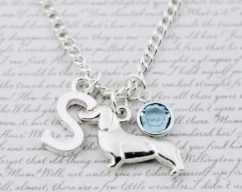May include: Silver necklace with a dachshund charm, a letter "S" pendant, and a small blue gemstone. The chain is a classic curb style, and the charms are detailed. The background is a blurred, handwritten script.