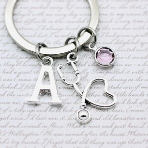 May include: Silver keychain with a charm of the letter A, a stethoscope, a heart, and a pink crystal. The keychain is on a white background with text.