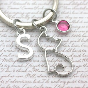 May include: A silver keychain with a letter 'S' charm, a small cat charm, and a pink rhinestone charm. The keychain is on a white background with cursive writing.