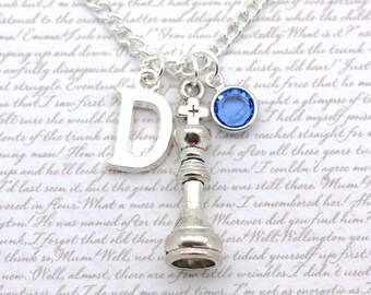 May include: Silver necklace with a charm of a chess king, a letter 'D' charm, and a light blue crystal charm. The necklace is on a white background with handwritten text.