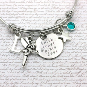 May include: A silver charm bracelet with a charm that reads "faith trust & pixie dust". The bracelet also features a silver fairy charm, a silver letter L charm, a silver star charm, and a turquoise blue crystal charm.